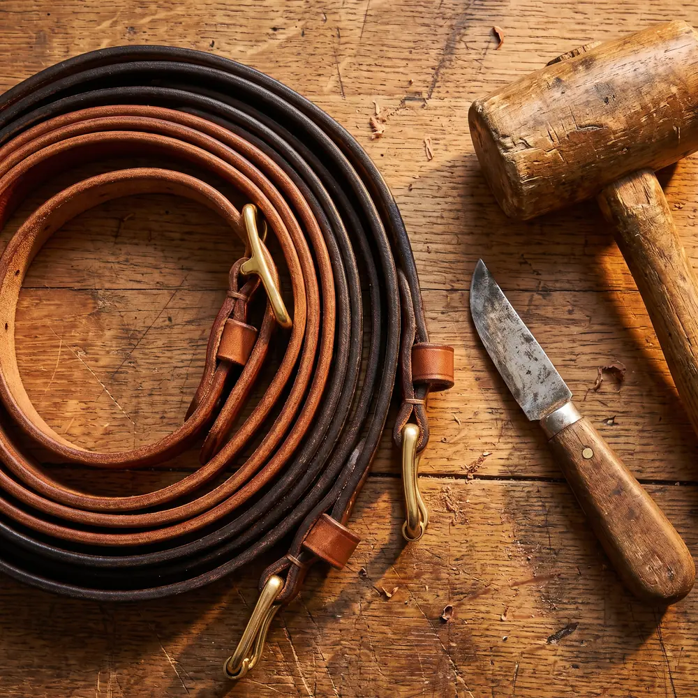 Coiled bridle leather straps and brass buckles on a worn workbench beside a cutting knife and mallet.