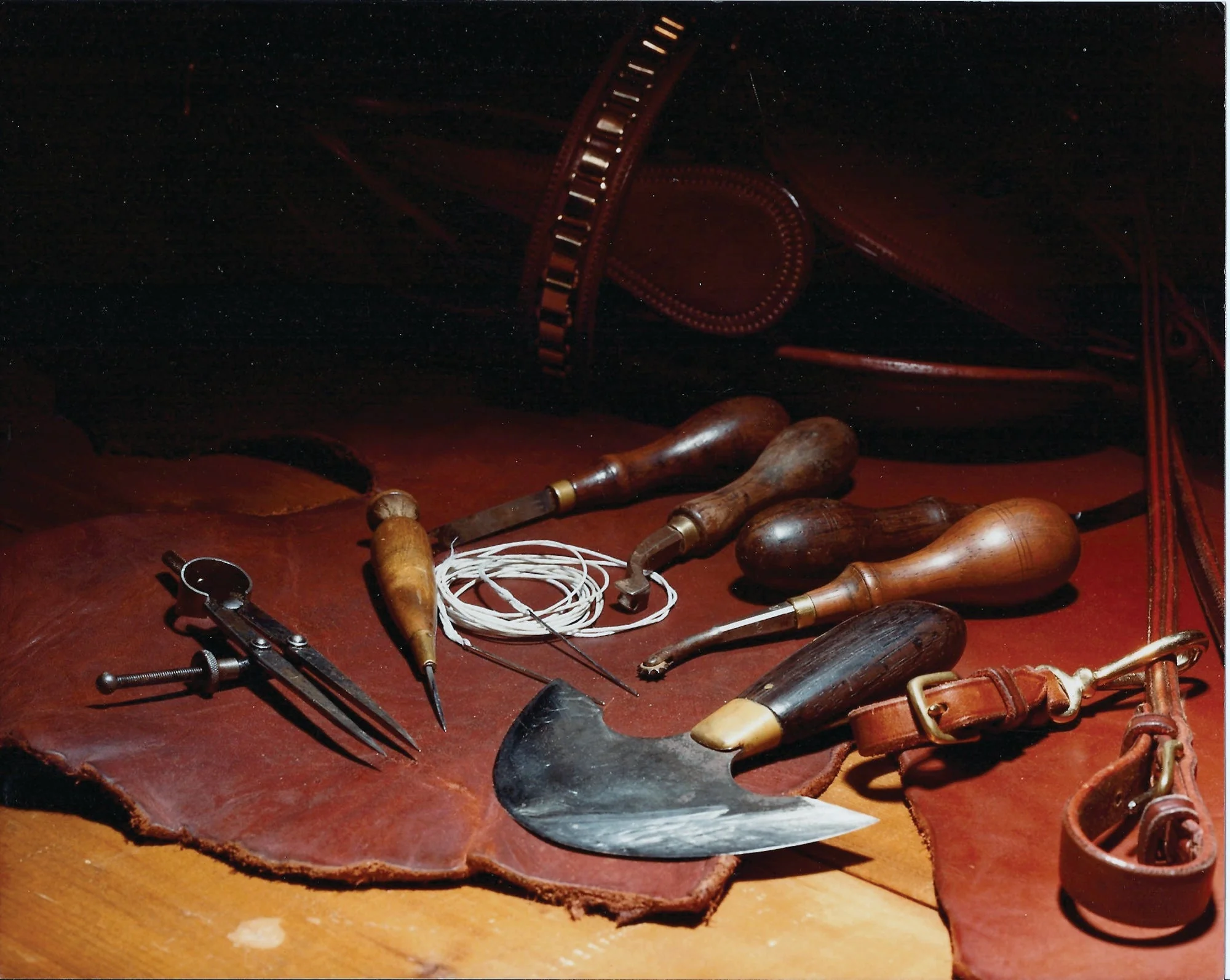 A leatherworker's hand tools laid across a cut hide on the bench: edge beveler, awls, waxed linen thread and a half-moon skiving knife.