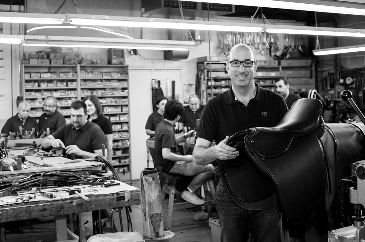 David Freedman holding a finished saddle in the Toronto workshop, with the bench team stitching and fitting hardware at their stations behind him.